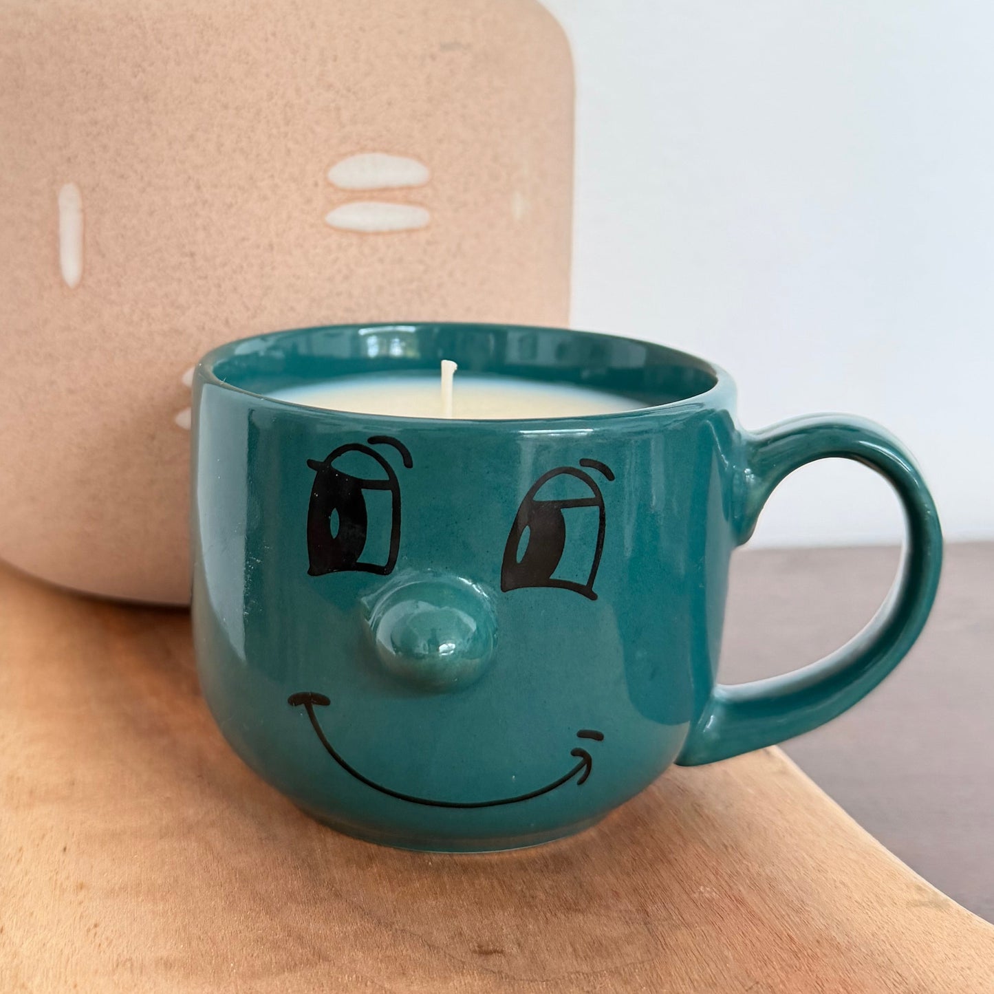 A teal ceramic mug candle with a hand-drawn smiling face sits on a wooden surface in front of a beige textured vase with green leaves.