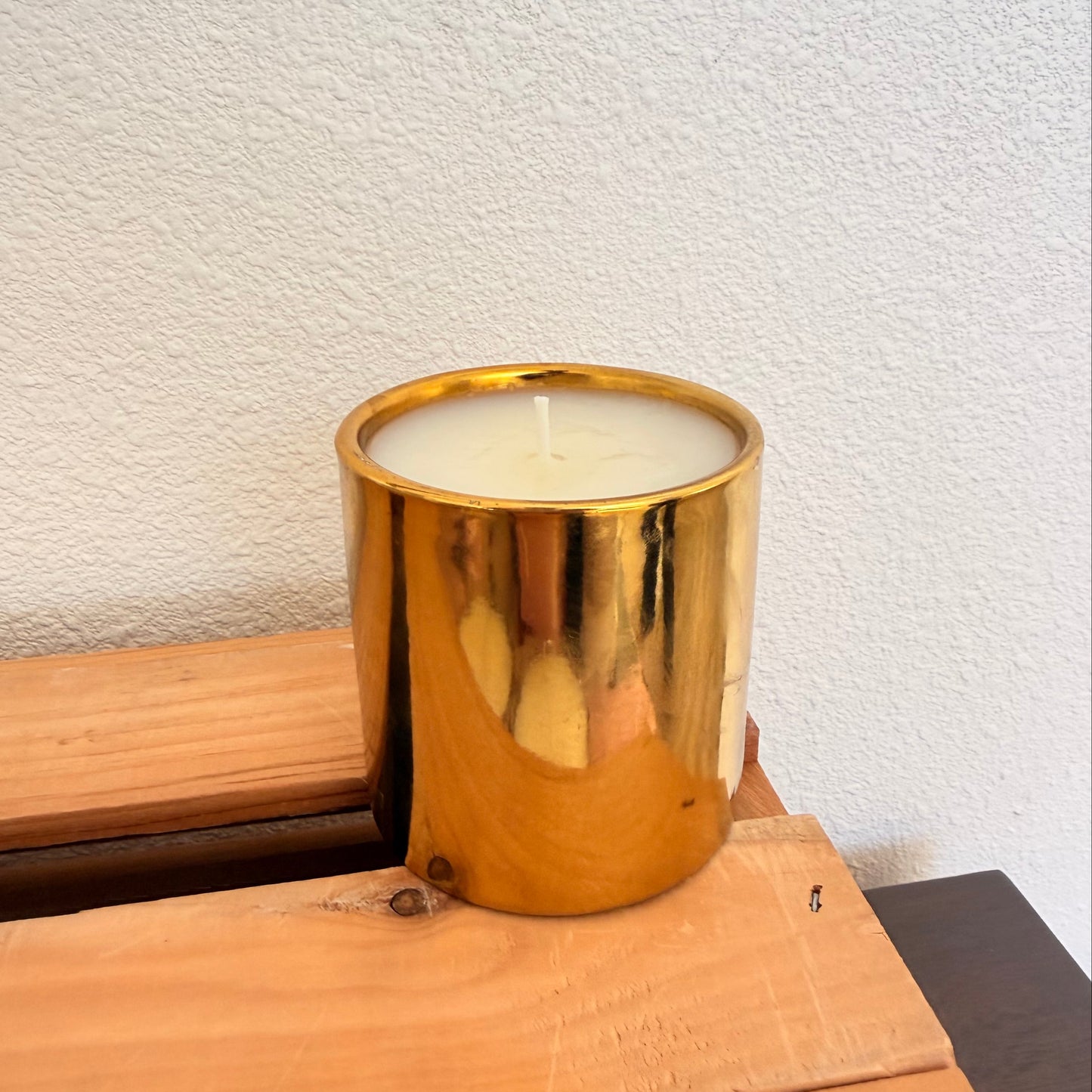 A gold metallic cylindrical candle holder containing white wax, resting on a light-colored wooden surface against a textured off-white wall.