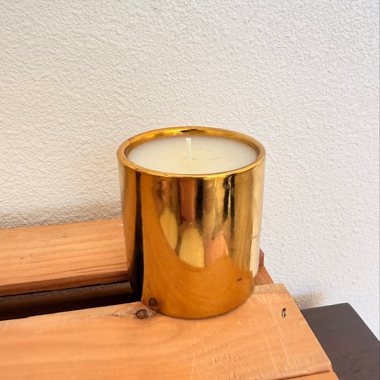 A gold metallic cylindrical candle holder containing white wax, resting on a light-colored wooden surface against a textured off-white wall.