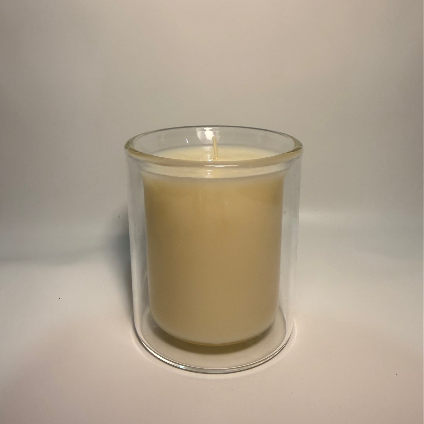 A single-wick candle in a clear glass jar with a double-walled glass design, filled with a light cream-colored wax, set against a plain light gray background.