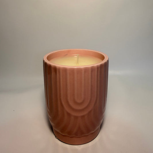 A single lit candle in a ribbed, terracotta-colored ceramic vessel with a cream-colored wax surface, set against a neutral light gray background.